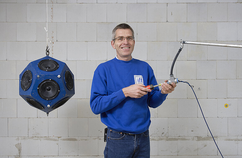 An test engineer looks into the camera, next to him are a microphone and a loudspeaker for acoustic tests