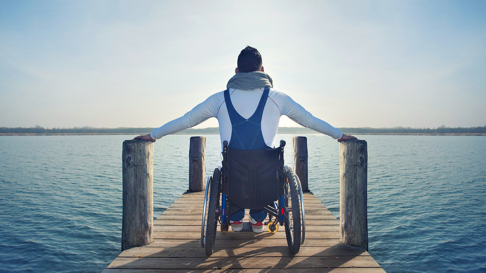 Auf einem Steg an einem See sitze ein Mann im Rollstuhl und berührt rechts und links die Pfosten. Das Bild steht symbolisch für den Bereich Barrierefreiheit