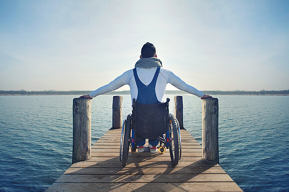 Das Foto zeigt einen Rollstuhlfahrer auf einem Steg am See (Quelle: © Jenny Sturm – stock.adobe.com)
