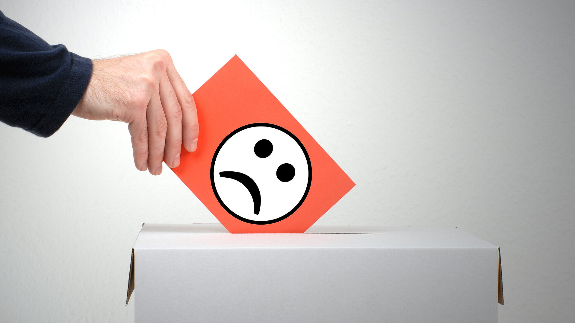 Hand wirft Zettel mit schlechtgelauntem Smiley in eine Wahl-Box