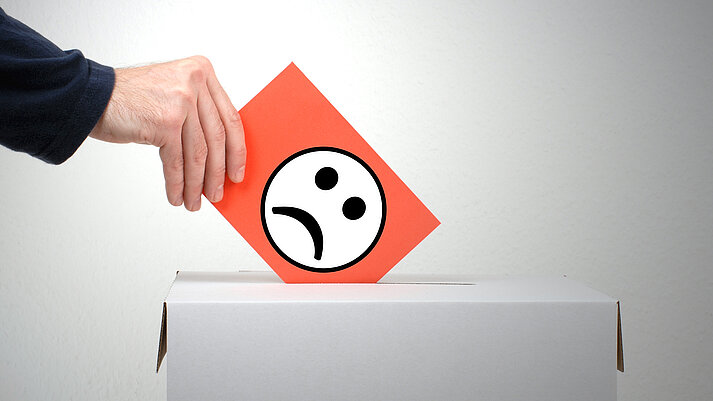 Hand wirft Zettel mit schlechtgelauntem Smiley in eine Wahl-Box