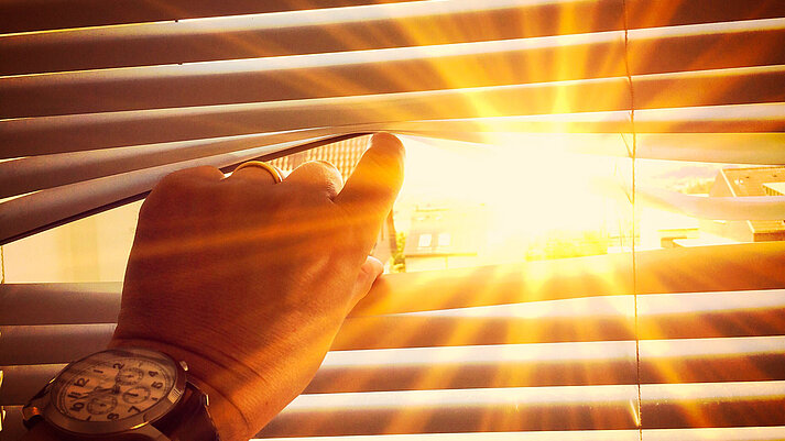 Eine Hand öffnet einen Spalt in einer Jalousie, durch das dahinterliegende Fenster scheint die Sonne