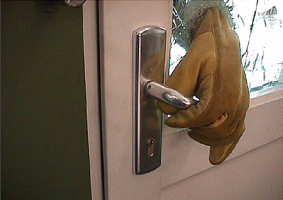 Foto der Hand eines Einbrechers, nachdem die Scheibe einer Tür zerschlagen wurde und jetzt mit der Klinke die Tür geöffnet wird.