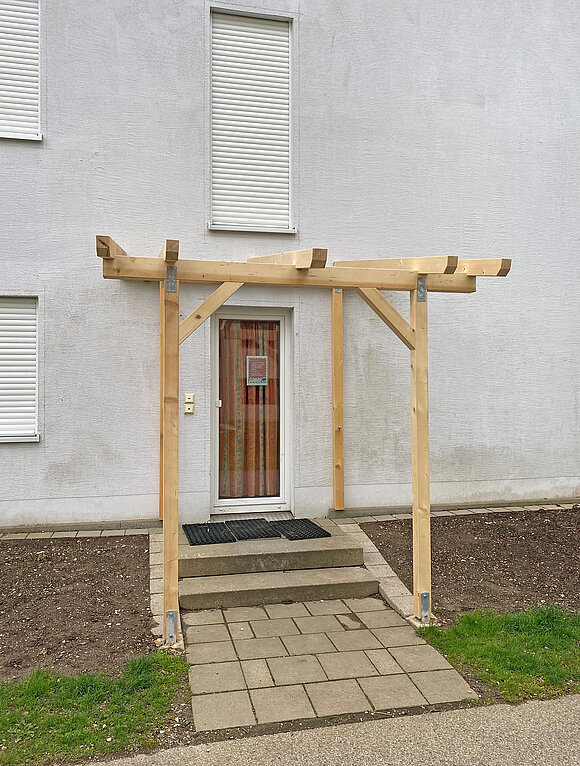 Das Schaubild zeigt eine Tür mit einem nachgerüsteten Vordach aus Holz. Nähere Informationen zur Darstellung erhalten Sie auf Anfrage unter +49 8031 261-2150.