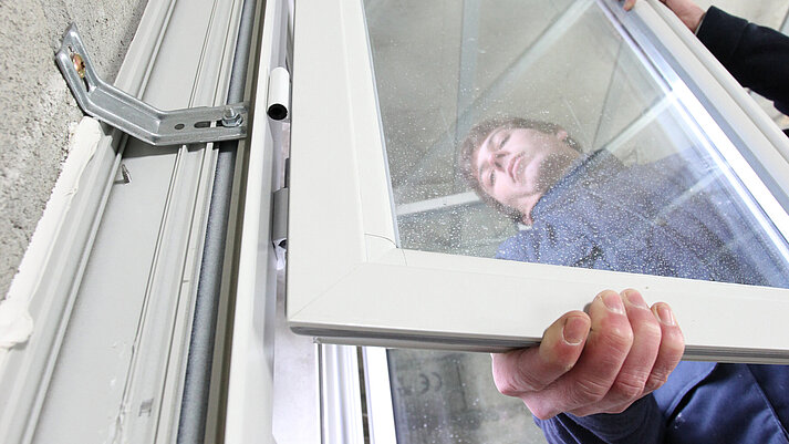 Ein Handwerker montiert vor einer Betonwand einen Flügel an einem Fensterelement. Das Bild steht symbolisch für den Bereich Montage