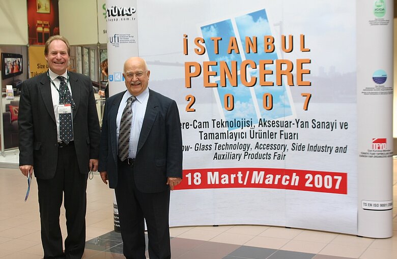 two men standing next to each other laughing in front of a large poster bearing the inscription “Istanbul Pencere 2007”