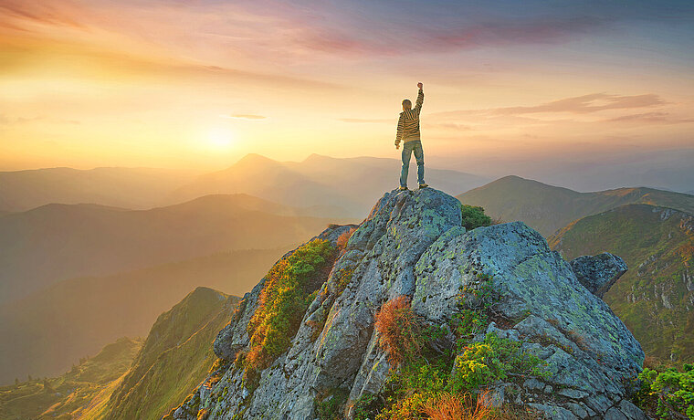 Person steht mit erhobener Faust auf einer Bergspitze im Sonnenaufgang 