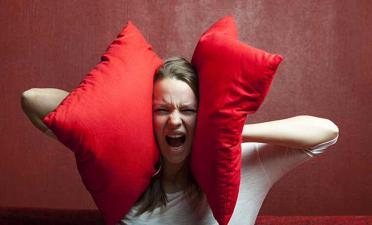 Eine schreiende Frau hält sich zwei rote Kissen an die Ohren. Das Bild steht symbolisch für Schallschutz