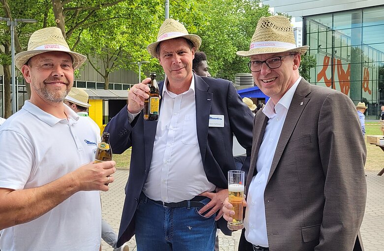 Das Foto zeigt drei Männer mit Strohhüten auf dem Kopf, die zusammen Bier trinken.