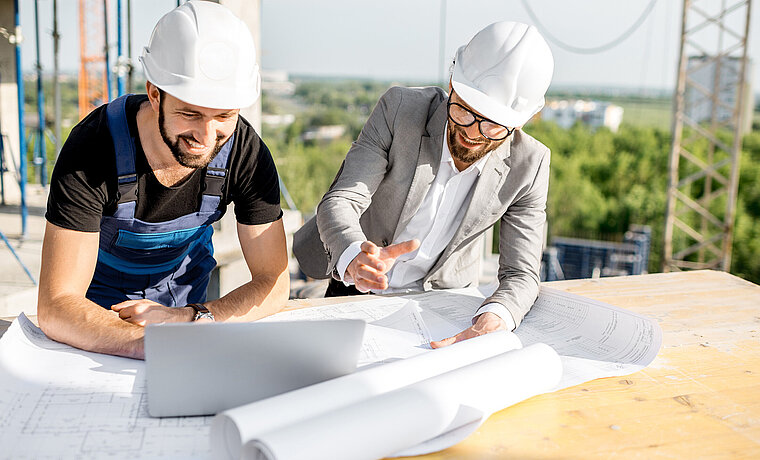 Zwei Männer mit Bauhelmen auf einer Baustelle stehen gebeugt über einem Bauplan