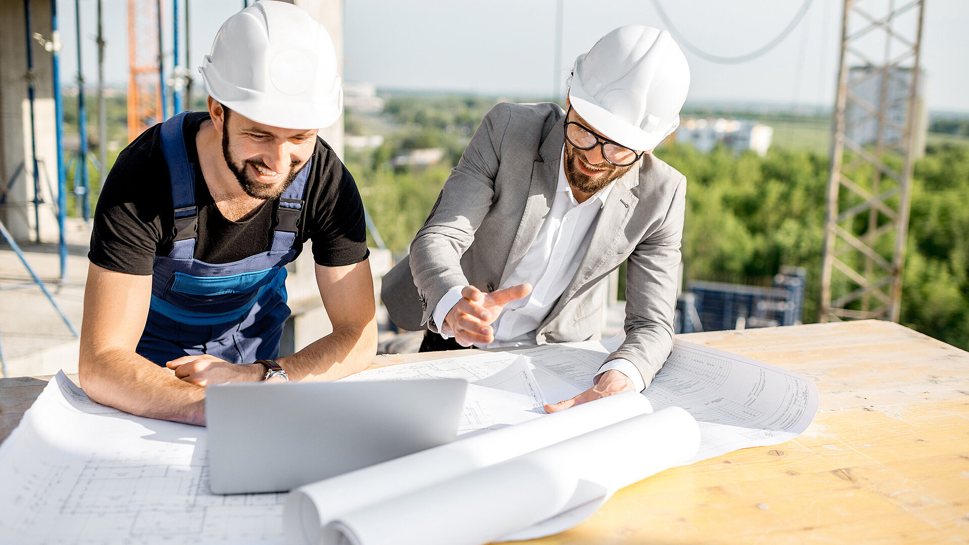 Zwei Männer mit Bauhelmen auf einer Baustelle stehen gebeugt über einem Bauplan