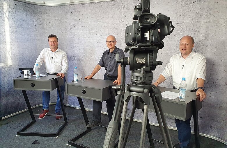 Drei Männer stehen vor der Kamera hinter drei Rednerpulte. 